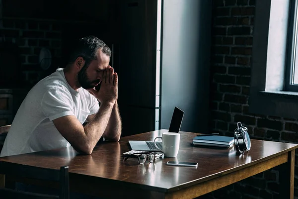 Depressed young man sitting on kitchen with laptop and leaning head on hands — Stock Photo