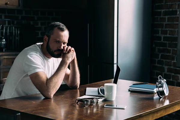 Depressed young man sitting on kitchen with laptop and looking away — Stock Photo