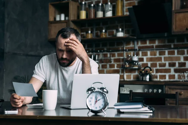 Joven deprimido mirando la tarjeta de fotos en la mano mientras está sentado en la cocina con el ordenador portátil y el reloj despertador - foto de stock