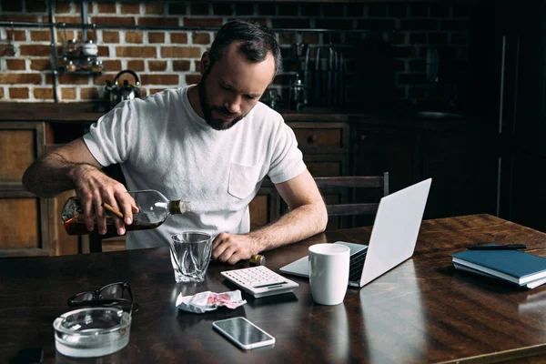 Alcoholic young man pouring whiskey in glass on kitchen — Stock Photo