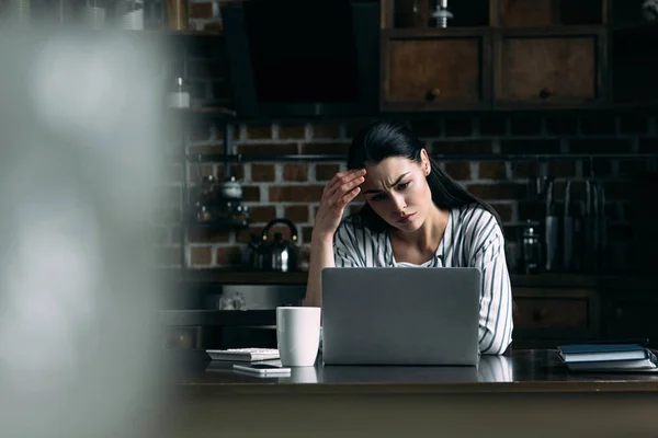 Jeune femme déprimée regardant écran d'ordinateur portable tout en étant assis sur la cuisine — Photo de stock