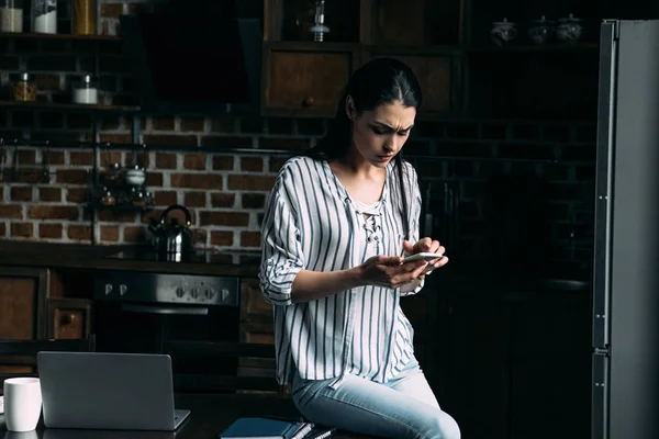 Sad young woman sitting on table at kitchen and using smartphone — Stock Photo