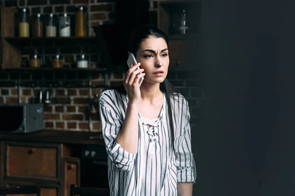 Sad young woman talking by phone and looking away on kitchen — Stock Photo