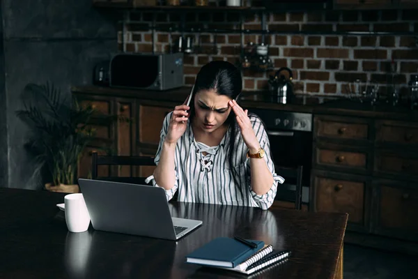 Mujer joven deprimida hablando por teléfono y mirando el ordenador portátil en la cocina - foto de stock