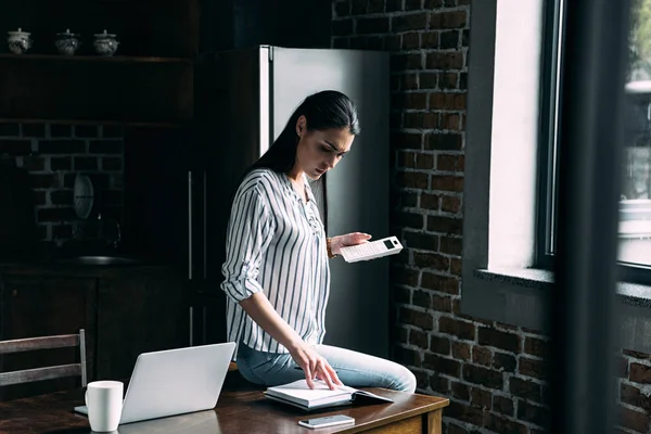 Triste joven con calculadora contando impuestos en la cocina en casa - foto de stock