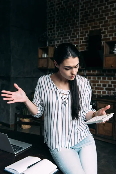 Bewildered young woman with calculator counting tax at home and shredding shoulders — Stock Photo