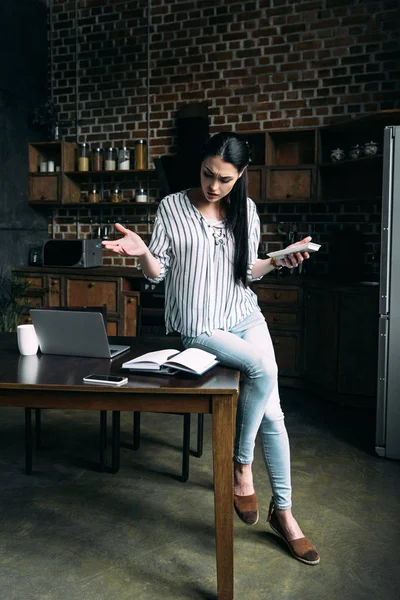 Растерянная молодая женщина с калькулятором подсчета налогов и глядя на ноутбук на кухне — стоковое фото