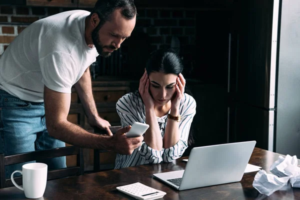 Depressed young woman sitting at table with laptop and crumpled papers while her angry husband showing her smartphone screen — Stock Photo