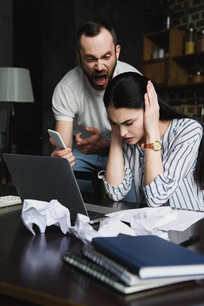 Mujer joven estresada sentada en la mesa con el ordenador portátil y los papeles arrugados mientras su marido le grita - foto de stock