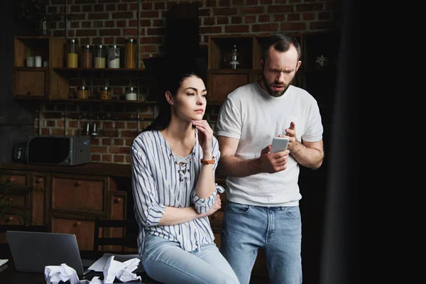 Mujer joven sentada en la mesa en la cocina mientras su marido mira el teléfono inteligente y grita - foto de stock