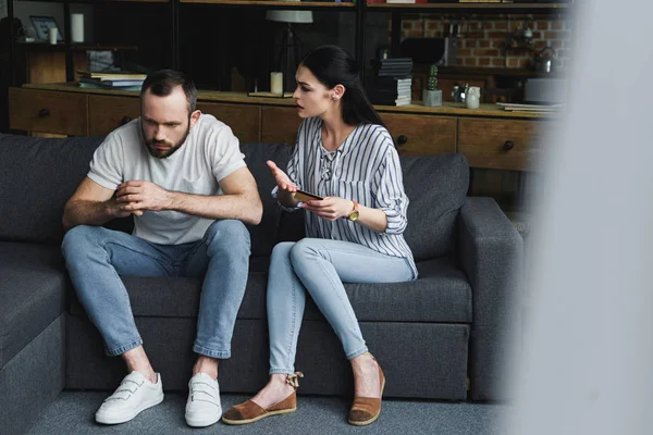 Sad young man sitting on sofa and looking away while his wife shouting at him and holding credit card — Stock Photo