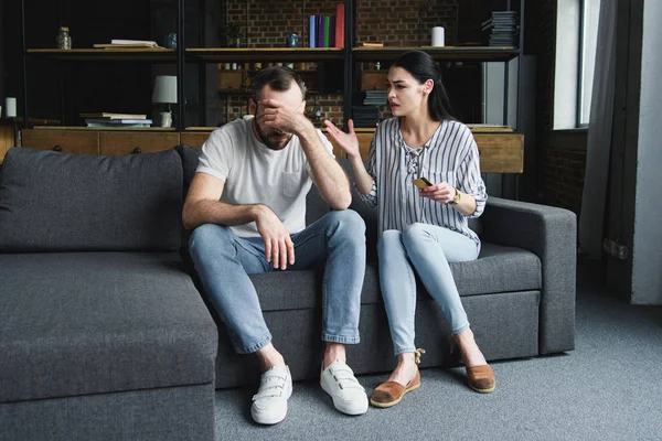 Triste joven sentado en el sofá y mirando hacia otro lado mientras su esposa le grita y sostiene la tarjeta de crédito - foto de stock