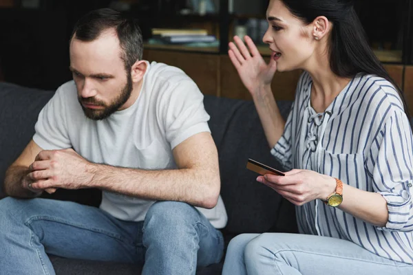 Deprimido joven sentado en el sofá y mirando hacia otro lado mientras su esposa le grita y sostiene la tarjeta de crédito - foto de stock