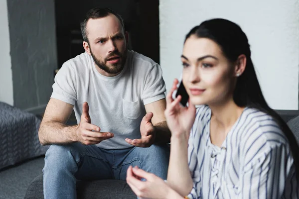 Angry man talking to his wife while she talking by phone on couch — Stock Photo