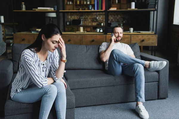 Femme triste assis sur le canapé tandis que son mari parle par téléphone brouillé sur le fond — Photo de stock