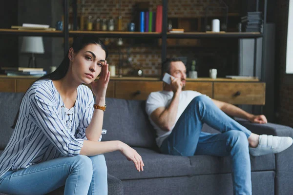 Depressed woman sitting on couch while her husband talking by phone blurred on background — Stock Photo