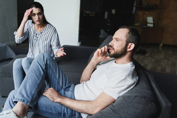 Femme folle parler à son mari alors qu'il parle par téléphone sur le canapé — Photo de stock