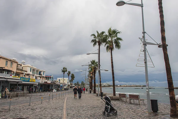 PAPHOS, CHIPRE - 31 DE MARZO DE 2020: carruaje de bebé cerca de la orilla del mar, personas y palmeras - foto de stock