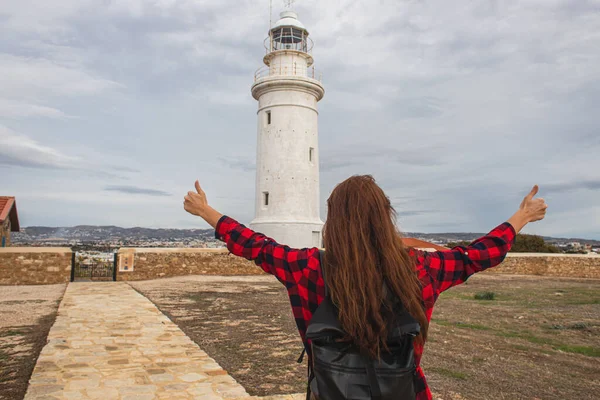 Vue arrière de la femme avec sac à dos montrant pouces vers le haut près du phare blanc — Photo de stock