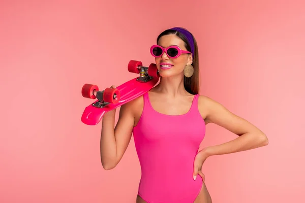 Vista frontale della donna in occhiali da sole con mano sul fianco sorridente e tenendo penny board isolato su rosa — Foto stock