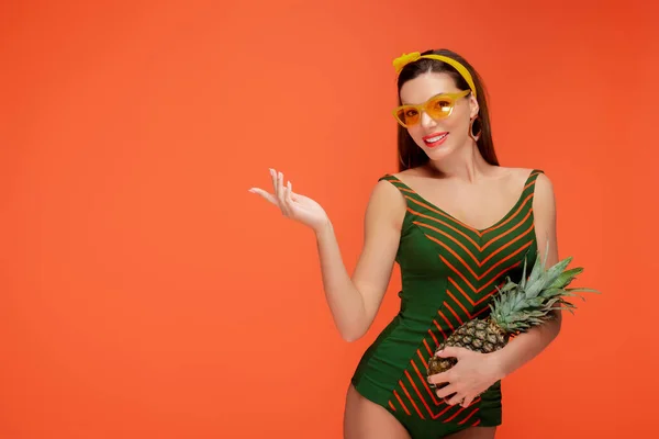 Vista frontale della donna con ananas che punta con mano, sorridente e guardando la fotocamera isolata su arancione — Foto stock