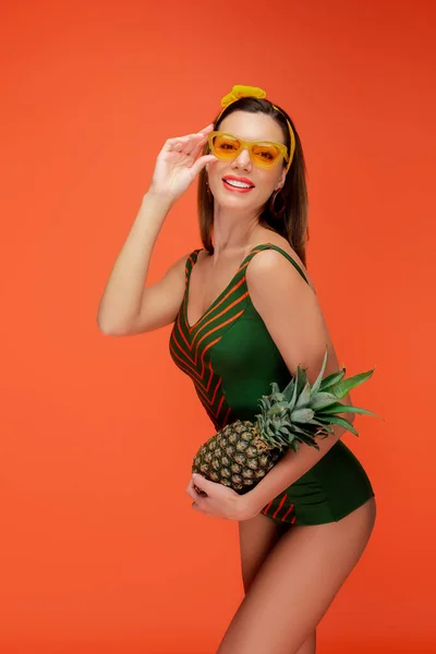 Mujer con gafas de sol sosteniendo piña sonriendo y mirando a la cámara aislada en naranja - foto de stock
