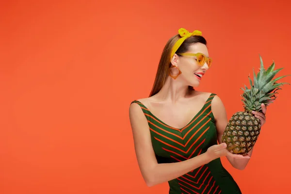 Mujer sonriendo y mirando a la piña aislada en naranja - foto de stock