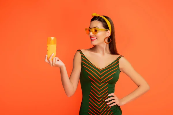 Vista frontal de la mujer con la mano en la cadera mirando botella de protector solar y sonriendo aislado en naranja - foto de stock