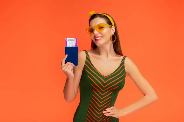 Vista frontal de la mujer con la mano en la cadera sonriendo y mostrando pasaporte con billete de avión aislado en naranja - foto de stock