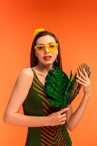 Mulher de óculos de sol segurando folhas verdes e olhando para a câmera isolada em laranja — Fotografia de Stock