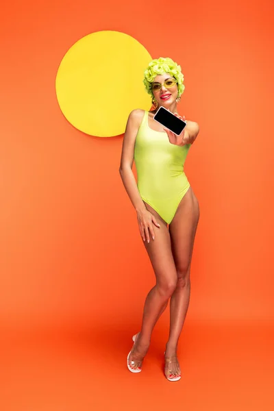 Женщина в шляпе с декоративными цветами смотрит в камеру, улыбается и показывает смартфон с желтым кругом сзади на оранжевом — стоковое фото