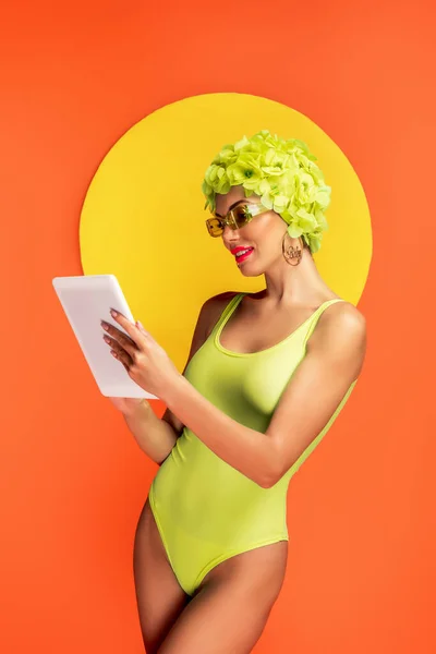 Frau mit Hut mit dekorativen Blumen blickt auf digitales Tablet mit gelbem Kreis dahinter auf orange — Stockfoto
