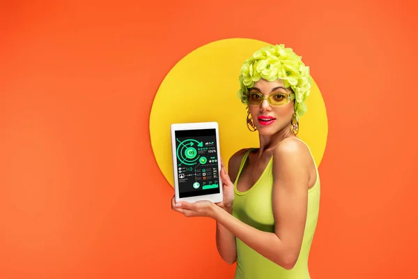 Donna in cappello con fiori decorativi che presenta tablet digitale con app e guardando la fotocamera con cerchio giallo dietro su arancione — Foto stock