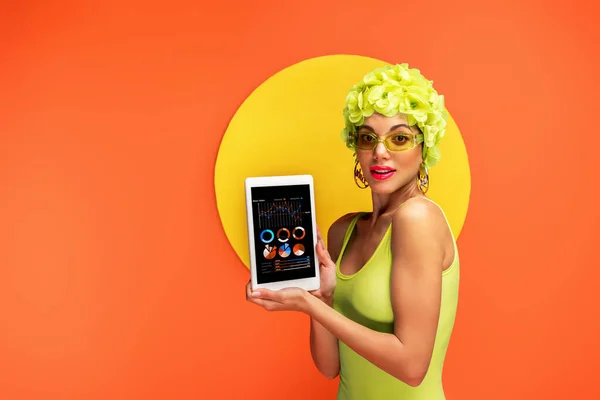 Chica en sombrero con flores decorativas que muestran tableta digital con aplicación y mirando a la cámara con círculo amarillo detrás en naranja - foto de stock