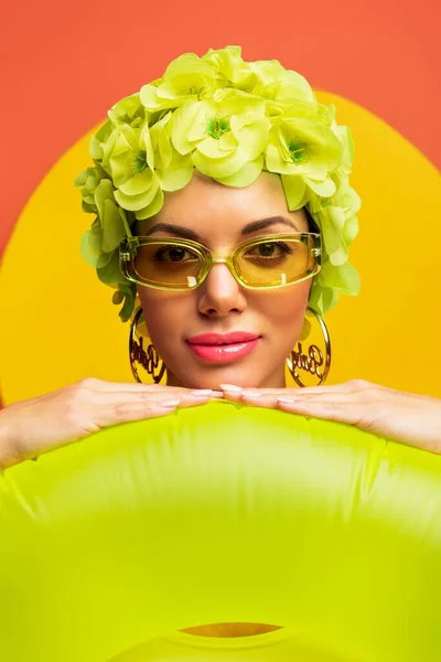 Ritratto di ragazza in cappello con fiori decorativi e occhiali da sole che sorride e mette le mani su anello di nuoto su giallo e arancione — Foto stock