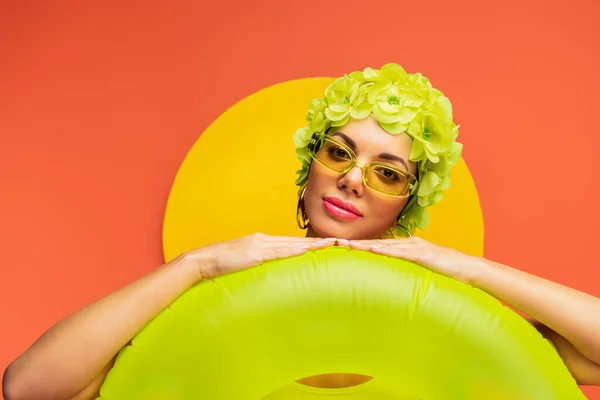 Portrait of girl in hat with decorative flowers and sunglasses putting hands on swim ring on yellow and orange — Stock Photo