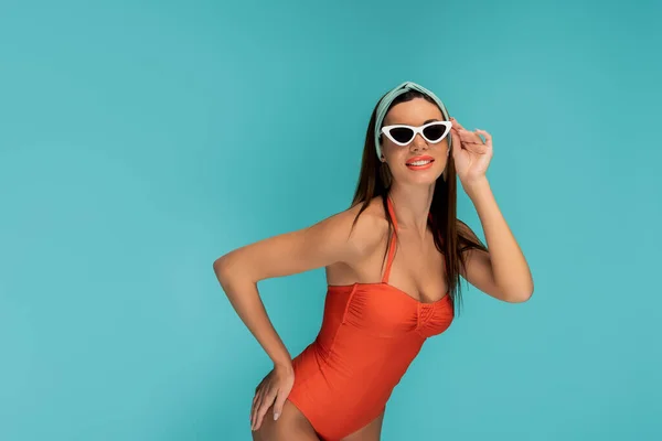 Donna con mano sull'anca toccando occhiali da sole e sorridendo isolata sul blu — Foto stock