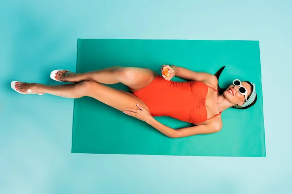 Вид сверху женщины с бутылкой солнцезащитного крема улыбающейся и лежащей на пляжном одеяле на голубом — стоковое фото