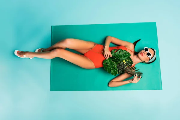 Вид зверху на жінку з зеленим листям, що лежить на пляжній ковдрі на синьому — стокове фото
