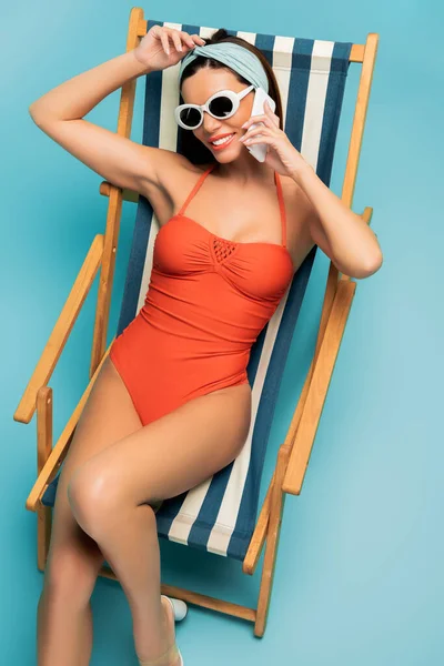 Visão de alto ângulo da mulher sorrindo, falando no smartphone e sentado na cadeira de praia em azul — Fotografia de Stock