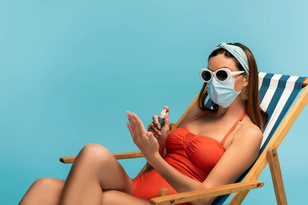 Frau mit Sonnenbrille und medizinischer Maske benutzt Händedesinfektionsmittel und sitzt auf Liegestuhl isoliert auf blauem Grund — Stockfoto