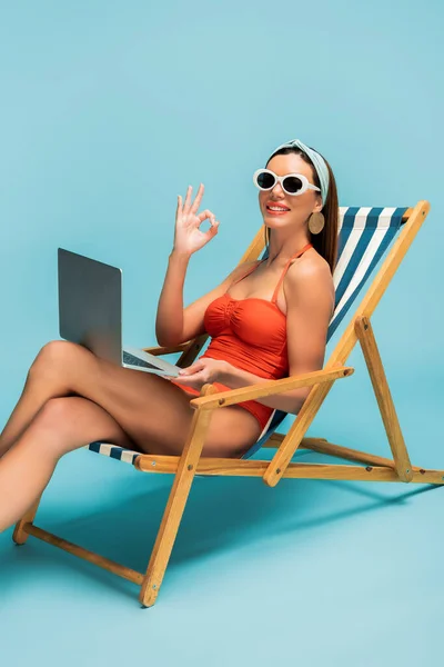 Freelancer com pernas cruzadas e laptop sorrindo e mostrando sinal ok na cadeira de praia em azul — Fotografia de Stock