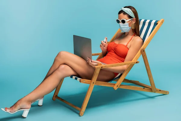 Freelancer in medical mask with laptop and bottle of hand sanitizer on deckchair on blue — Stock Photo