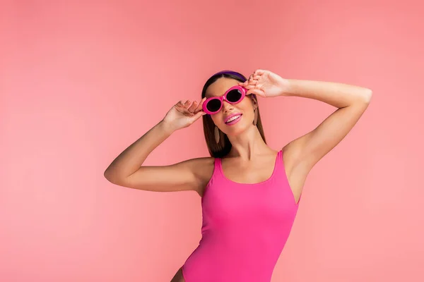 Mujer sonriendo y tocando gafas de sol aisladas en rosa - foto de stock
