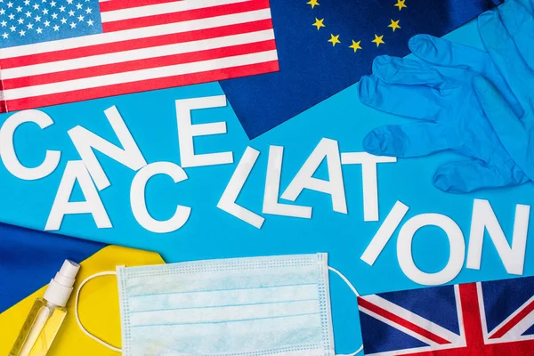 Top view of lettering cancellation near medical mask, latex gloves and flags of countries on blue surface — Stock Photo