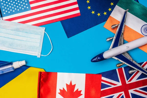 Vista superior do avião de brinquedo perto de máscara médica e bandeiras de países no fundo azul — Fotografia de Stock