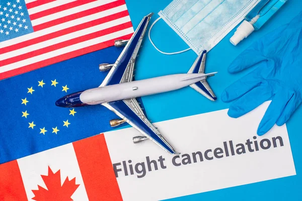 Top view of toy plane, card with flight cancellation lettering near medical mask and flags of countries on blue surface — Stock Photo