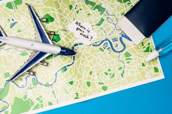 Top view of toy plane near speech bubble with where is your mask lettering near passport and bottle of hand sanitizer on map on blue surface — Stock Photo