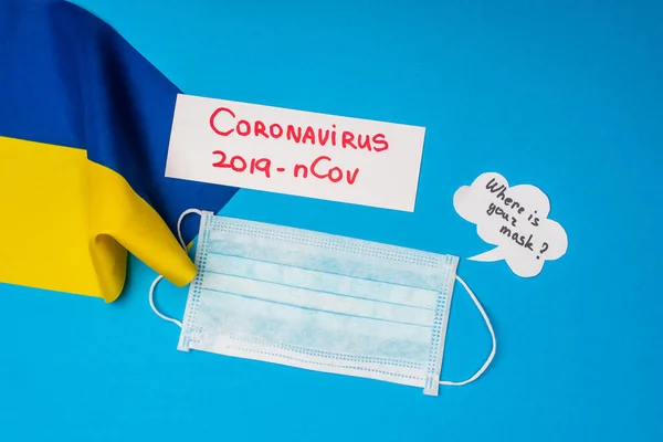 Vista superior de la tarjeta con letras coronavirus 2019-nCov, máscara médica y dónde está su máscara en la burbuja del habla con la bandera de Ucrania en la superficie azul - foto de stock