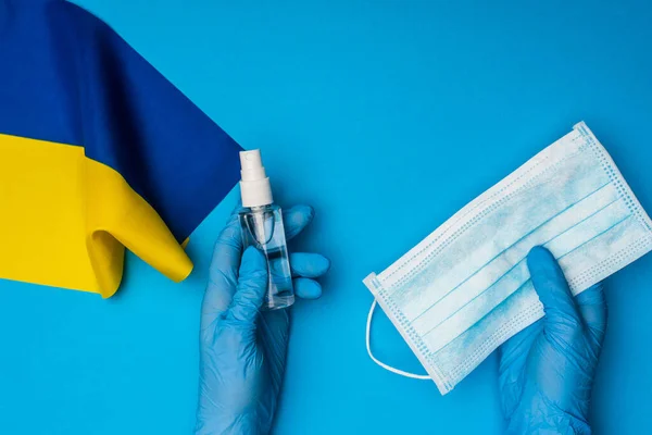 Vista superior del médico sosteniendo la botella con desinfectante de manos y máscara médica cerca de la bandera de Ucrania sobre fondo azul - foto de stock